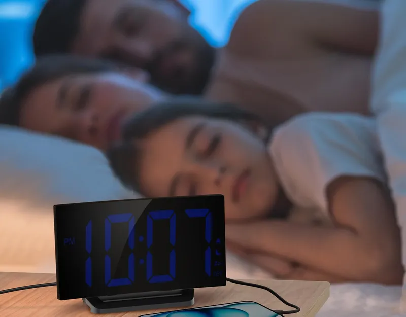 Curved alarm clock on a nightstand, screen dimmed, family asleep behind it